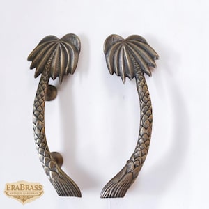Könnte beinhalten: Zwei bronzefarbene Türgriffe in Form von Palmen. Die Griffe sind gebogen, mit einem Palmenwedel oben und einem strukturierten Stamm. Sie sind auf einer weißen Oberfläche montiert. Das Logo "ERABRASS ANTIQUE HARDWARE" ist sichtbar.