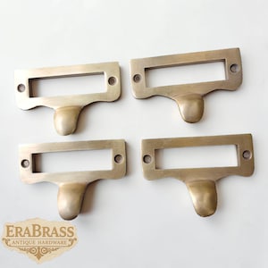 May include: Four antique brass drawer pulls, each with a rectangular label holder and a rounded pull. The pulls have a warm, aged brass finish and are arranged on a white surface. The EraBrass logo is visible in the bottom left corner.