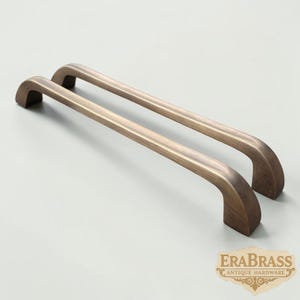 Peut inclure: Deux poignées de placard assorties avec une finition en laiton antique brossé. Chaque poignée présente un design arqué lisse avec des extrémités arrondies et une base de montage rectangulaire. Le logo EraBrass Antique Hardware est visible.