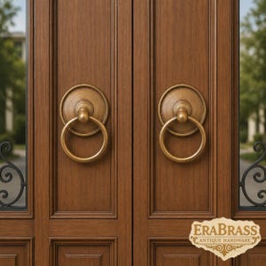 May include: Two circular brass door knockers with a ring design mounted on a pair of dark brown wooden doors. Each knocker features a round backplate and a swinging ring. The text ERABRASS ANTIQUE HARDWARE is visible in the bottom right corner.
