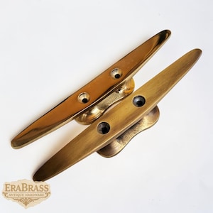 Puede incluir: Dos cornamusas de latón, una pulida y otra con pátina, sobre un fondo blanco. Cada cornamusa tiene una base central con dos agujeros y una parte superior larga y cónica. El logotipo "ERA BRASS ANTIQUE HARDWARE" es visible.