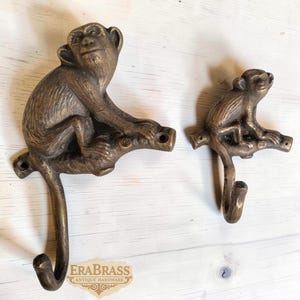 Puede incluir: Dos ganchos con forma de mono de color bronce. Los monos están posados en una rama, con un gancho que se extiende hacia abajo. Los ganchos son probablemente para colgar objetos. La imagen incluye el texto "ERABRASS Antique Hardware".