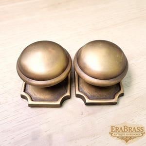 May include: Two matching brass door knobs with a smooth, rounded top and a square base featuring decorative cut corners. The metal has a muted, aged finish with subtle variations in tone. The text EraBrass Antique Hardware is visible.