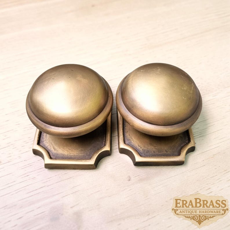 May include: Two matching brass door knobs with a smooth, rounded top and a square base featuring decorative cut corners. The metal has a muted, aged finish with subtle variations in tone. The text EraBrass Antique Hardware is visible.