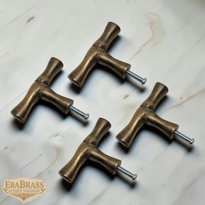 Può includere: Quattro pomelli per mobili antichi a forma di T in ottone con viti argentate. Le maniglie hanno un design a forma di bambù e sono disposte su una superficie di colore chiaro. Il logo Era Brass è visibile in basso a sinistra.