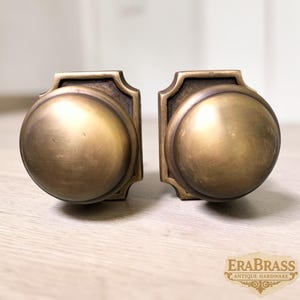 May include: Two matching round door knobs made of aged brass with a smooth, domed surface and a square-shaped backplate featuring beveled edges. The text EraBrass Antique Hardware is visible in the bottom right corner of the image.