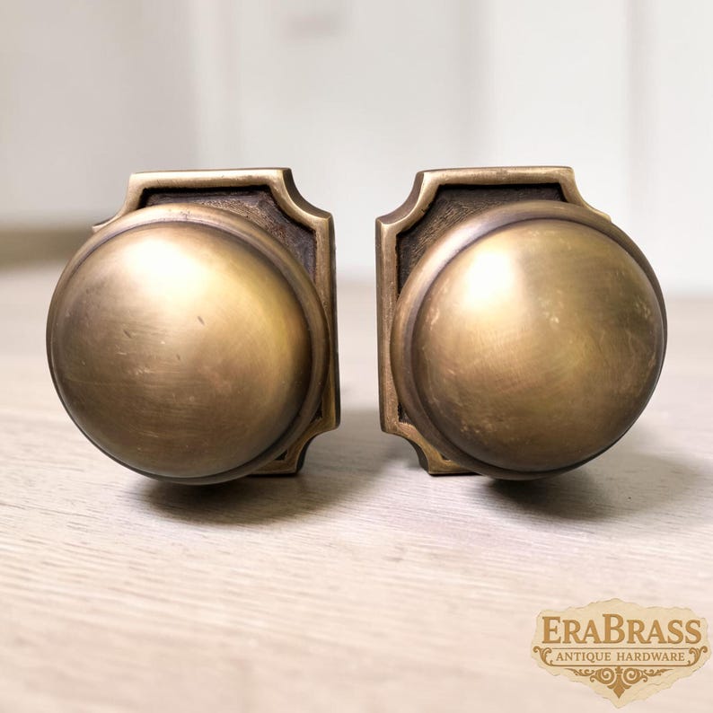 May include: Two matching round door knobs made of aged brass with a smooth, domed surface and a square-shaped backplate featuring beveled edges. The text EraBrass Antique Hardware is visible in the bottom right corner of the image.