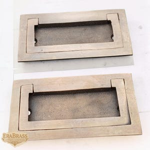 Puede incluir: Dos tiradores empotrados de latón antiguo. Cada tirador rectangular tiene un área de agarre empotrada y está hecho de metal marrón claro. Los tiradores son probablemente para un armario o cajón. El logotipo "ERABRASS ANTIQUE HARDWARE" es visible.