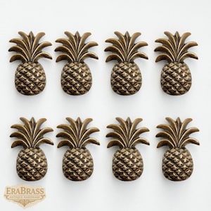 May include: Eight antique brass pineapple cabinet knobs arranged in two rows of four. Each knob features a detailed pineapple design with textured skin and a leafy crown. The knobs are a warm, metallic color, perfect for adding a tropical touch to furniture.
