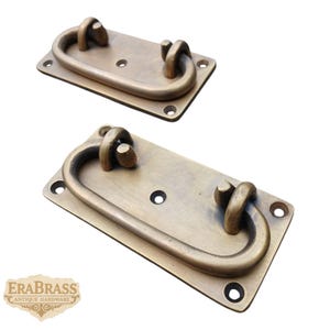 May include: Two antique brass drawer pulls. Each pull has a rectangular base with rounded corners and an oval handle. The pulls are a warm, aged brass color, with visible screw holes for mounting. The logo "ERABRASS ANTIQUE HARDWARE" is in the lower left corner.