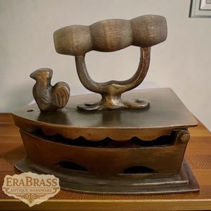 May include: Antique brass iron with a wooden handle and a decorative rooster figurine. The iron has a rectangular base and a curved handle. The text "ERABRASS ANTIQUE HARDWARE" is visible on the bottom left.