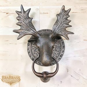 May include: Bronze moose head door knocker with large antlers and a circular backplate. The knocker has a ring-shaped handle and is mounted on a white wooden door. The logo "ERABRASS ANTIQUE HARDWARE" is visible in the lower left corner.