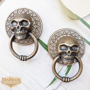 Puede incluir: Dos aldabas de puerta de calavera de latón antiguo. Cada aldaba presenta un diseño detallado de calavera con un asa de anillo. La calavera está montada sobre una placa circular con un patrón geométrico grabado. Se muestran sobre una superficie blanca.
