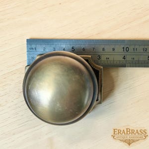 May include: A round brass door knob with a smooth, matte finish, positioned next to a metal ruler showing a diameter of approximately 2.5 inches. The hardware features a square base plate and is marked with the text EraBrass Antique Hardware.
