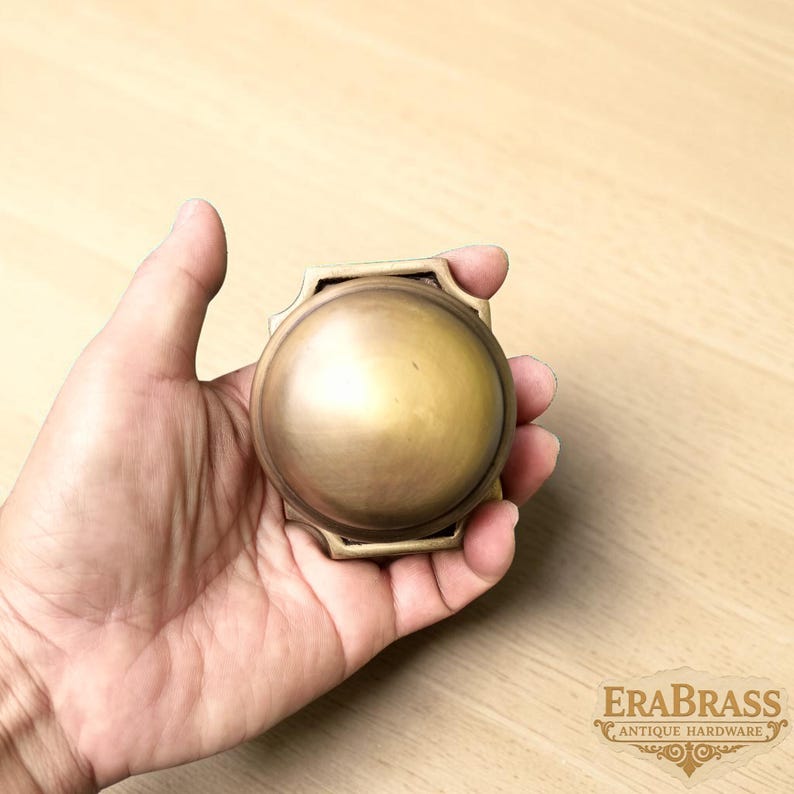May include: A circular brass door knob with a smooth, domed surface and a square-shaped base plate. The metal has a muted, brushed finish. The text EraBrass Antique Hardware is visible in the bottom right corner of the image.