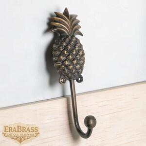 Puede incluir: Un gancho de pared decorativo con forma de piña, de color bronce. El gancho es de metal y tiene un diseño detallado de piña. Está montado sobre una superficie de madera clara y una pared blanca.