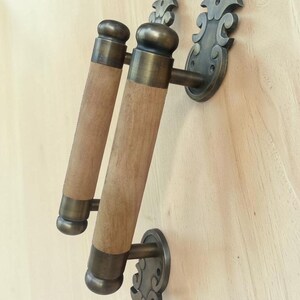 May include: A door handle featuring two cylindrical wooden grips accented with dark brass end caps and rounded finials. The handle is mounted to a surface using two ornate, fleur-de-lis inspired brass plates with visible screw holes.