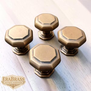 May include: Four antique brass cabinet knobs with an octagonal design. The knobs have a warm, aged finish and are arranged on a light-colored wooden surface. The knobs are from EraBrass Antique Hardware.