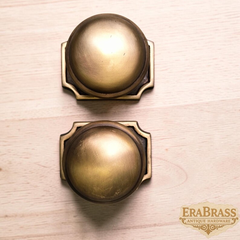 May include: Two round brass door knobs with a smooth, matte finish. Each knob is mounted on a rectangular backplate with decorative notched corners. The text "ERABRASS ANTIQUE HARDWARE" is visible in the bottom right corner of the image.