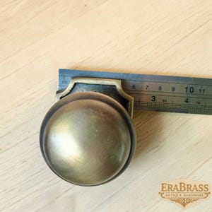May include: A round brass door knob with a dark, aged finish and a decorative backplate. A metal ruler is positioned against the knob, showing a width of approximately 2.5 inches. The knob has a smooth, domed surface and a metallic sheen.