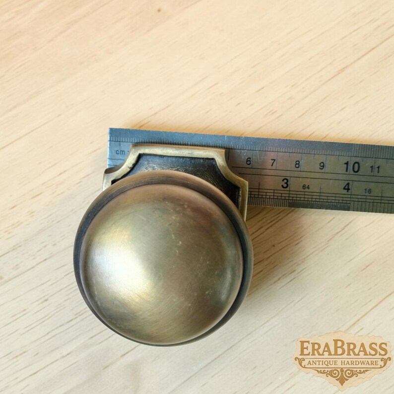 May include: A round brass door knob with a dark, aged finish and a decorative backplate. A metal ruler is positioned against the knob, showing a width of approximately 2.5 inches. The knob has a smooth, domed surface and a metallic sheen.