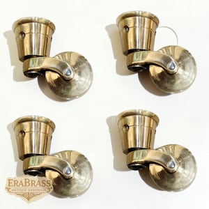 May include: Four antique brass casters, each with a cup-shaped top and a circular wheel. The casters are a warm gold colour, designed for furniture. The EraBrass logo is visible in the lower left corner.
