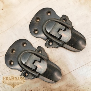 Puede incluir: Dos cierres de cofre antiguos de color bronce. Cada cierre tiene un cuerpo redondeado con un asa curvada y con bisagras y una placa de montaje con múltiples orificios para tornillos. Los cierres se muestran sobre una superficie de madera clara.