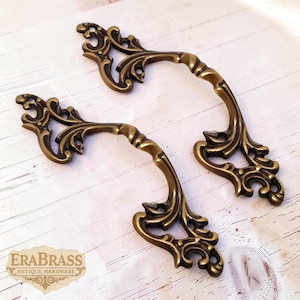 May include: Two antique brass drawer pulls with ornate, swirling floral designs. The pulls have a curved handle and decorative end pieces. The image includes the text "EraBrass Antique Hardware".