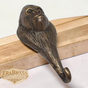 Peut inclure: Crochet en forme de gorille de couleur bronze. Le crochet présente des détails de fourrure et des traits du visage. Le crochet est monté sur une surface en bois. Le logo "ERABRASS ANTIQUE HARDWARE" est visible.
