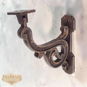 Può includere: Staffa da parete antica color ottone con un design a volute ornate. La staffa presenta una parte superiore quadrata e un braccio curvo con volute decorative. La piastra di montaggio rettangolare ha una parte superiore decorativa. Il logo "EraBrass Antique Hardware" è visibile.