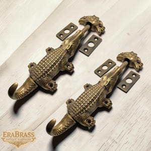 May include: Two antique brass alligator-shaped hooks with matching mounting plates. The hooks are detailed with textured scales and a curved tail. The alligator's head is at the top, and the hook extends from the tail. The logo "ERABRASS ANTIQUE HARDWARE" is at the bottom.
