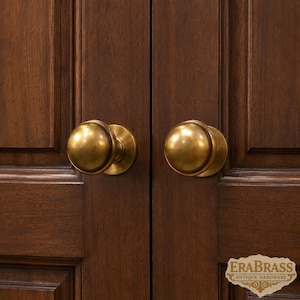 Puede incluir: Dos pomos de puerta redondos de metal en tono latón con un acabado liso y pulido, montados en puertas de armario de madera de color marrón oscuro. Una pequeña etiqueta decorativa de color crema en la esquina inferior derecha muestra el texto ERABRASS ANTIQUE HARDWARE.