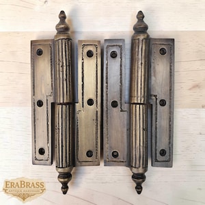 May include: Two antique brass hinges with decorative vertical lines and finial tops. Each hinge has a rectangular plate with screw holes. The hinges are displayed on a light wood surface. The logo "ERABRASS ANTIQUE HARDWARE" is visible.