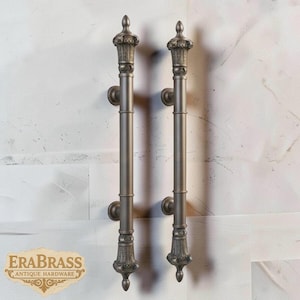 May include: Two antique bronze door pulls with ornate detailing. Each pull features a decorative top, a long cylindrical body, and a detailed base. The pulls are mounted on a white wall. The EraBrass logo is visible in the lower left corner.