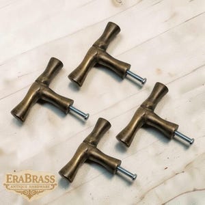 Könnte beinhalten: Vier antike T-förmige Schrankgriffe aus Messing mit silbernen Schrauben, angeordnet auf einer hellen Holzoberfläche. Die Griffe haben ein rustikales, gealtertes Aussehen. Das Logo "EraBrass Antique Hardware" ist unten links sichtbar.