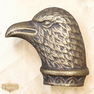 Może przedstawiać: Brązowa rzeźba głowy orła z szczegółowymi teksturami piór. Dziób orła jest lekko otwarty, a oko skupione. Rzeźba ma ozdobny pas u podstawy. Widoczne jest logo Era Brass.