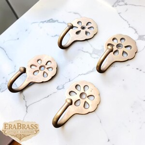 Peut inclure: Quatre crochets en laiton antique avec un motif floral. Chaque crochet a un bras incurvé et une plaque arrière décorative en forme de fleur. Les crochets sont disposés sur une surface en marbre blanc et gris. Le logo "EraBrass Antique Hardware" est visible.