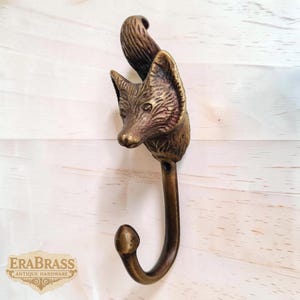 Op de afbeelding: Een bronzen vossenkop wandhaak. De vossenkop is gedetailleerd met getextureerde vacht en heeft een gebogen haak. De haak is gemonteerd op een lichtgekleurd houten oppervlak. De woorden "EraBrass Antique Hardware" staan linksonder.