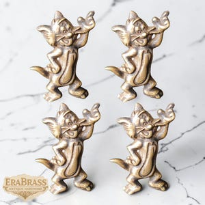 May include: Four antique brass cabinet pulls in the shape of a cartoon cat. The pulls are detailed with a smiling face, outstretched arms, and a tail. The pulls are made of brass and have a vintage finish. The logo "ERABRASS ANTIQUE HARDWARE" is in the bottom left corner.