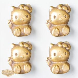 May include: Four brass cabinet knobs in the shape of a seated cartoon cat with a bow. The knobs are a warm gold color and have a detailed, antique finish. The knobs are arranged in a square pattern.
