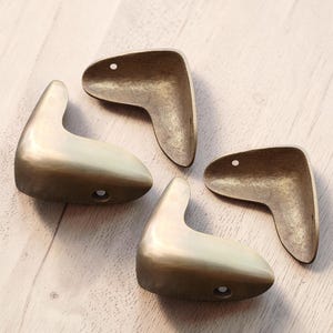 May include: Four antique brass-toned corner brackets, each with a curved design and a small hole. The brackets are arranged on a light-colored wooden surface, showcasing their aged appearance and unique shape. These are likely for furniture or decorative use.