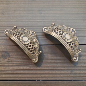 May include: A pair of antique brass drawer pulls with an ornate, openwork design. Each pull has a curved shape with a central medallion and decorative scrollwork. The pulls are set against a wooden background.