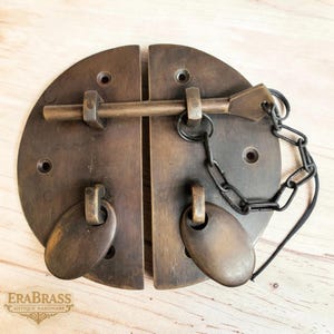May include: Antique brass cabinet latch with a circular design. The latch features a sliding bar, two oval pulls, and a chain. The hardware is a warm, aged bronze color, with visible screw holes. The brand name "ERABRASS" is visible.