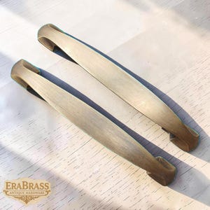 May include: Two antique brass cabinet pulls with a curved, elongated design. The pulls have a light, brushed finish and are set against a light-coloured surface. The logo "ERA BRASS ANTIQUE HARDWARE" is visible in the bottom left corner.