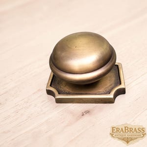 May include: A brass cabinet knob featuring a smooth, rounded dome shape attached to a square base with decorative notched corners. The metal has a warm, aged finish with visible dark patina accents. The text ERABRASS ANTIQUE HARDWARE is visible.