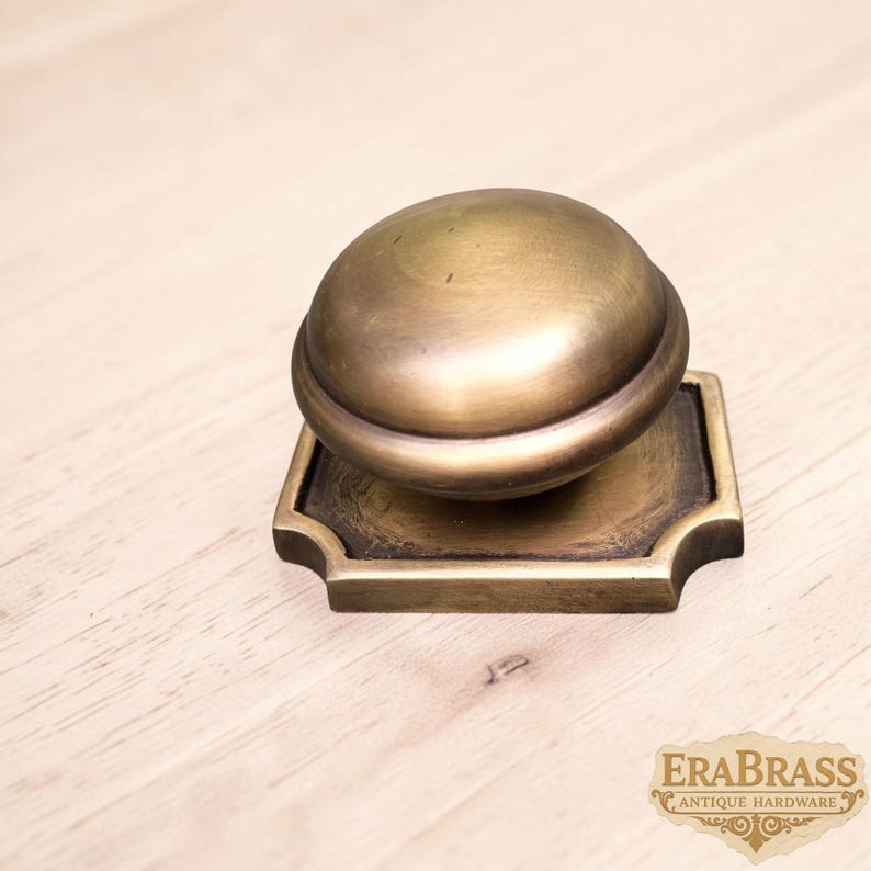 May include: A brass cabinet knob featuring a smooth, rounded dome shape attached to a square base with decorative notched corners. The metal has a warm, aged finish with visible dark patina accents. The text ERABRASS ANTIQUE HARDWARE is visible.