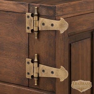 Puede incluir: Dos bisagras en T de latón de estilo antiguo montadas en un armario de madera marrón oscuro. Cada bisagra presenta un remate decorativo, una placa base rectangular con cuatro orificios para tornillos y un brazo largo. El texto ERABRASS ANTIQUE HARDWARE es visible.