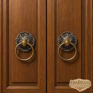May include: Two antique brass ring pulls with ornate circular backplates featuring intricate floral scrollwork, mounted on a pair of vertical wood cabinet doors. The bottom right corner displays the text ERABRASS ANTIQUE HARDWARE.