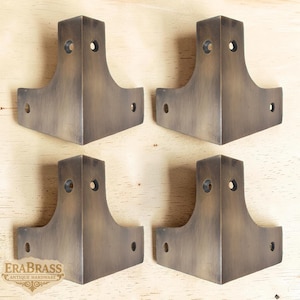 May include: Four antique brass corner brackets. Each bracket has a right-angle design with three pre-drilled holes. The brackets are a warm, aged brass color, and are displayed on a light wood surface. The EraBrass logo is visible in the lower left corner.