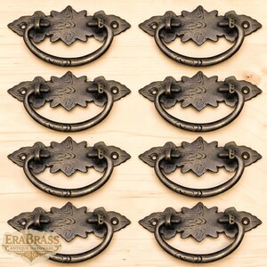 May include: Six antique bronze drawer pulls are displayed. Each pull has a decorative backplate with a floral design and a curved handle. The pulls are made of metal and have a vintage aesthetic. The brand name "ERABRASS" is visible on the bottom left.